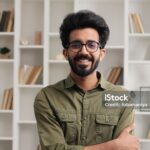 Gorgeous portrait of attractive confident Indian man with toothy smile looking at camera in modern living room. Businessman posing in casual stylish look in home office