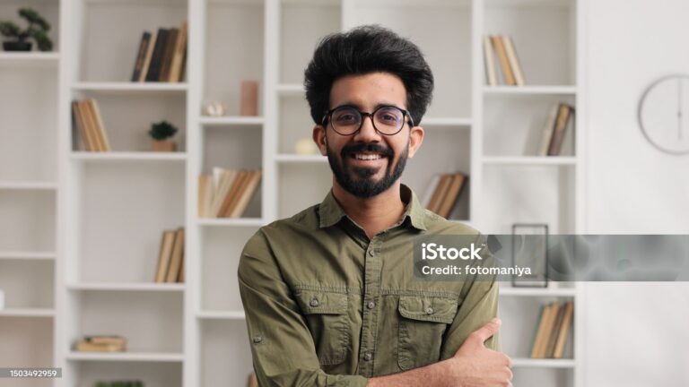 Gorgeous portrait of attractive confident Indian man with toothy smile looking at camera in modern living room. Businessman posing in casual stylish look in home office